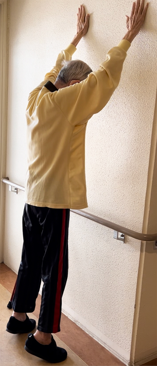 70歳代男性 壁に向かって両側腕を上げ、背伸びしている姿